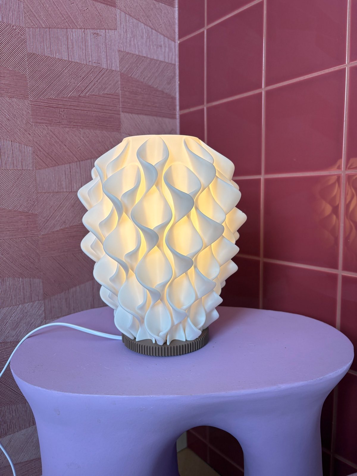 Coral tafellamp aan: warm licht schijnt door het golvende reliëf, op een lila bijzettafel voor roze tegels